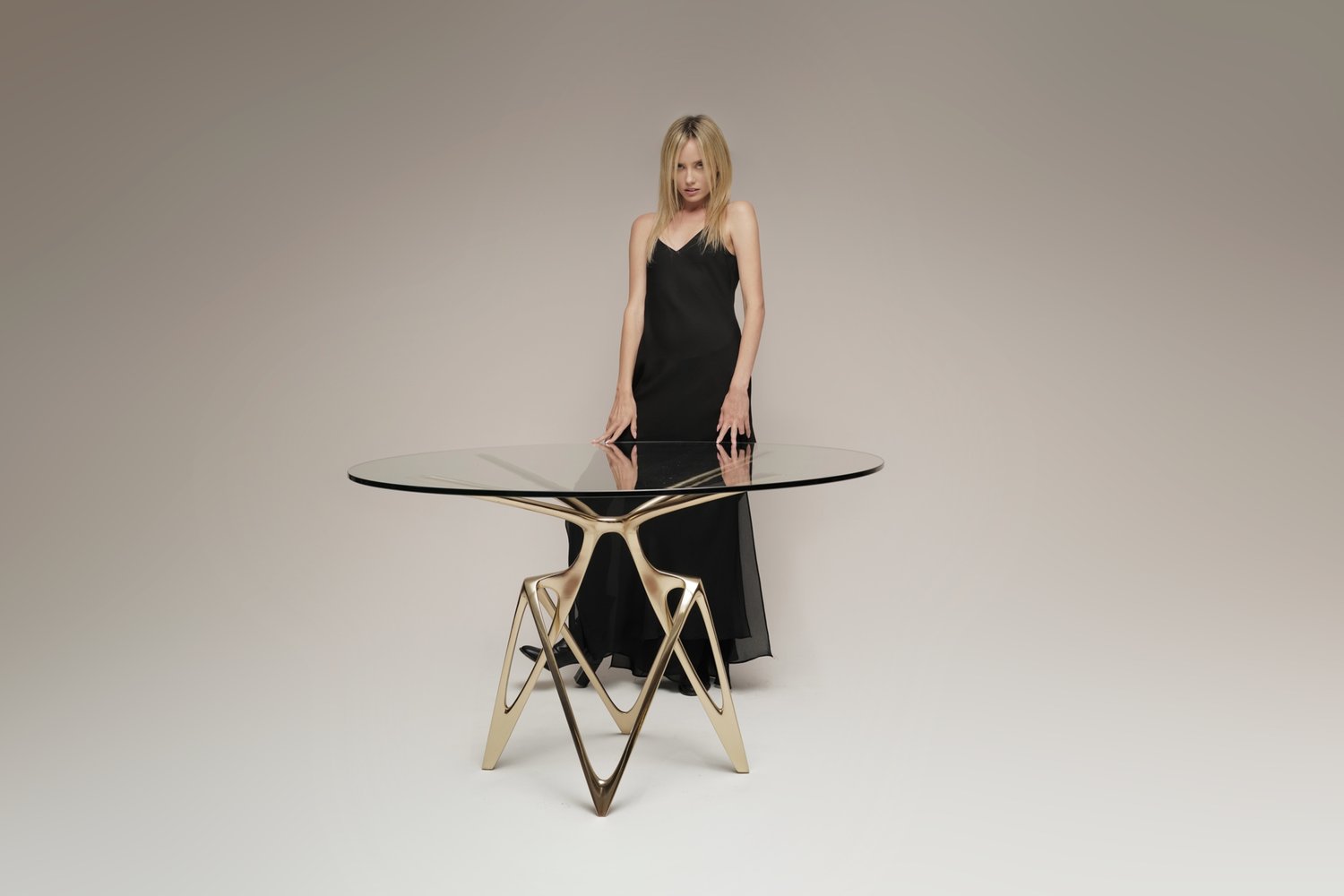 A woman in a black dress standing beside the Tronic Dining Table, featuring a sleek glass top and a unique geometric gold base.